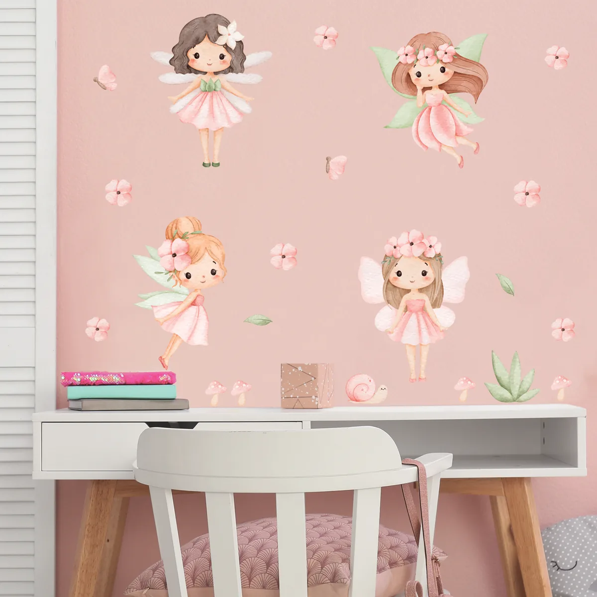 Parede rosa de quarto infantil decorada com adesivos de fadas aquareladas, flores e borboletas, com mesa branca e cadeira à frente